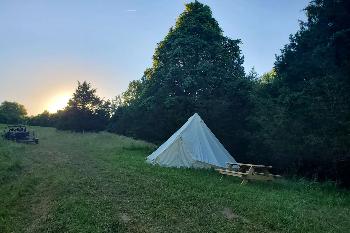 A canvas bell tent and picnic table in a mown field at sunset, with treeline behind